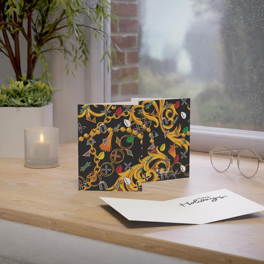 May include: Two folded greeting cards with a black background and gold baroque-style design featuring colorful chains, tassels, and other decorative elements.  The cards are displayed on a wooden surface, next to a lit candle and eyeglasses. A blank white greeting card is also shown, with the words 'Happy Holidays' written in elegant script.