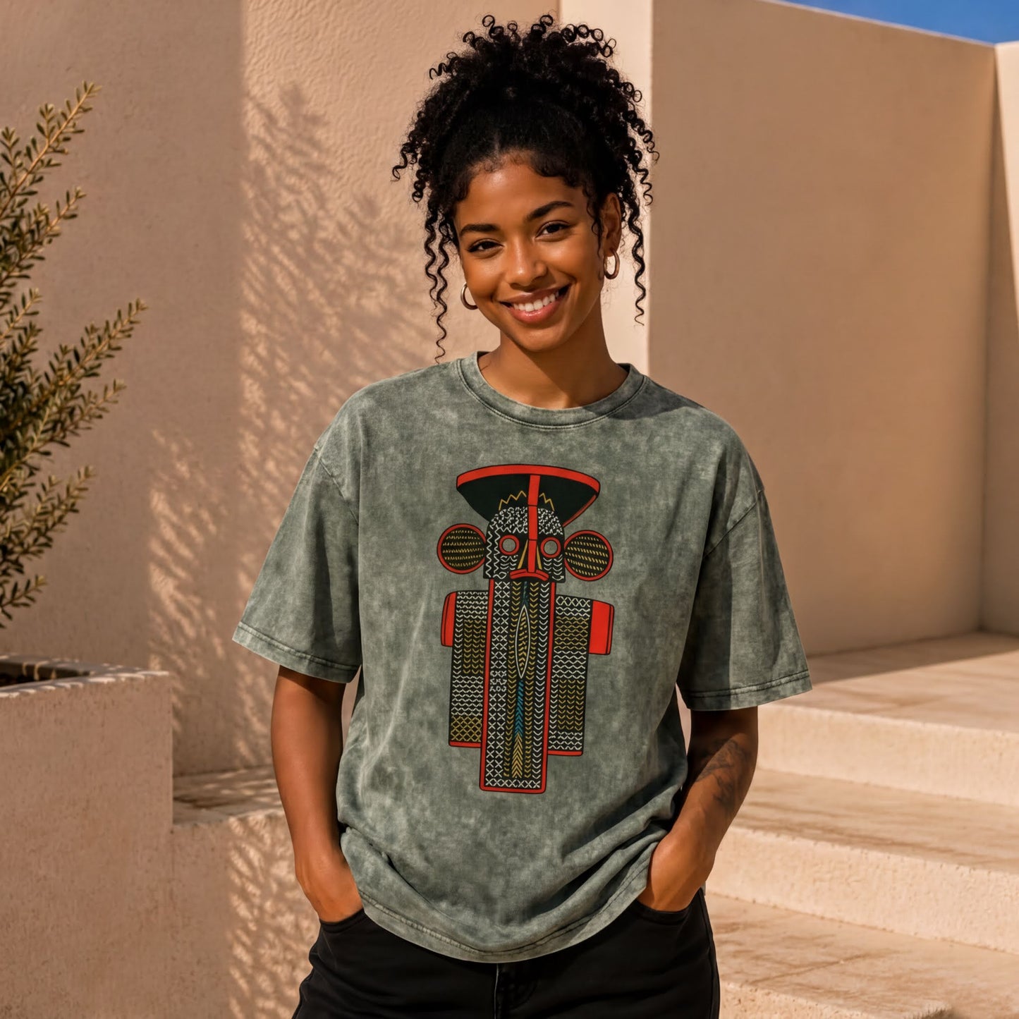 A young woman with curly hair is smiling and posing in front of a beige wall with a plant. She is wearing a gray t-shirt with a graphic design on it.
