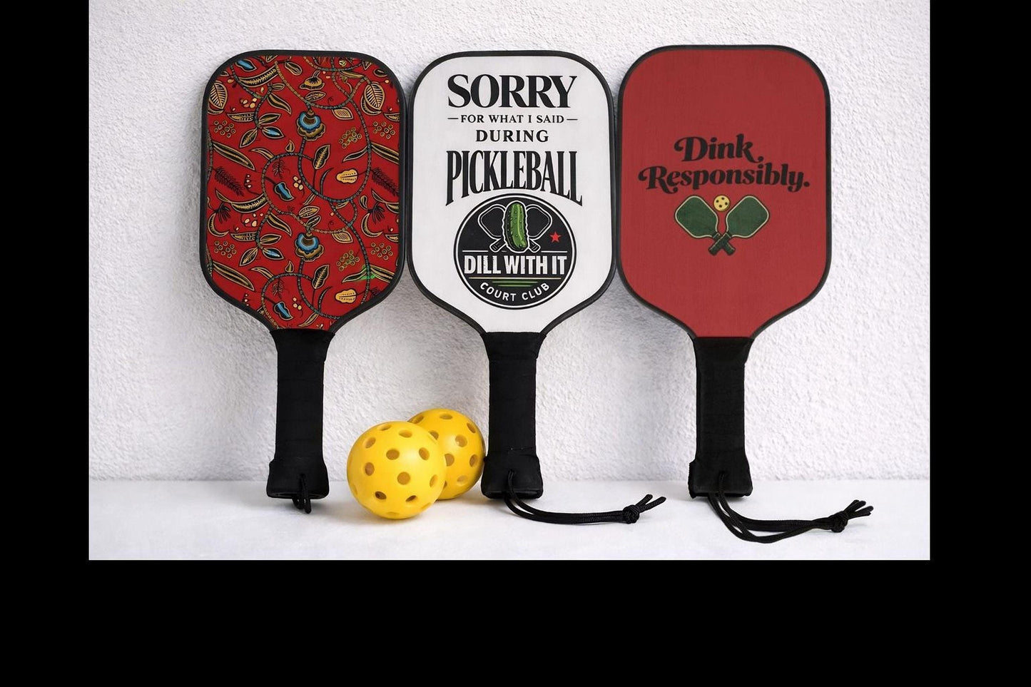 Three colorful paddleboards with different designs and text are displayed against a white wall, accompanied by two yellow balls.
