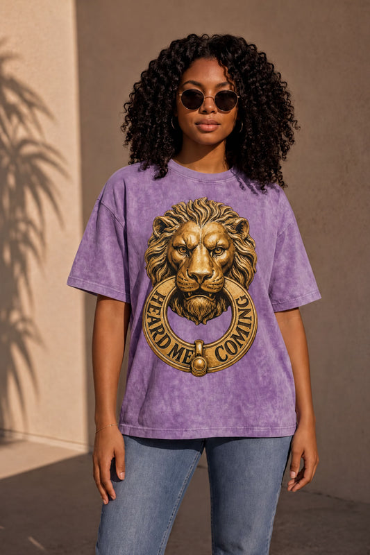 A young woman with curly hair wearing a purple t-shirt with a lion design and the text "HEARD ME COMING" is standing in front of a wall.