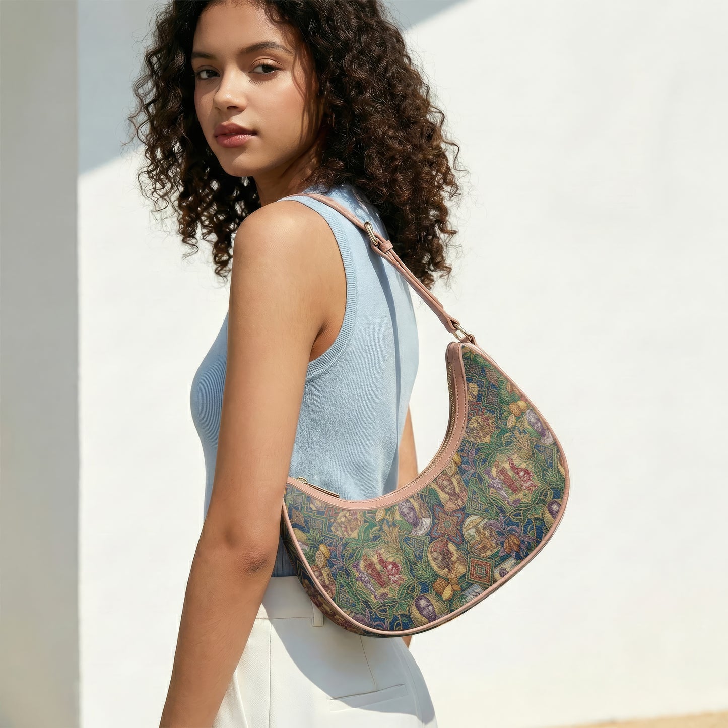 A young woman with curly hair is standing against a white wall, wearing a blue sleeveless top and carrying a colorful purse with a floral pattern.
