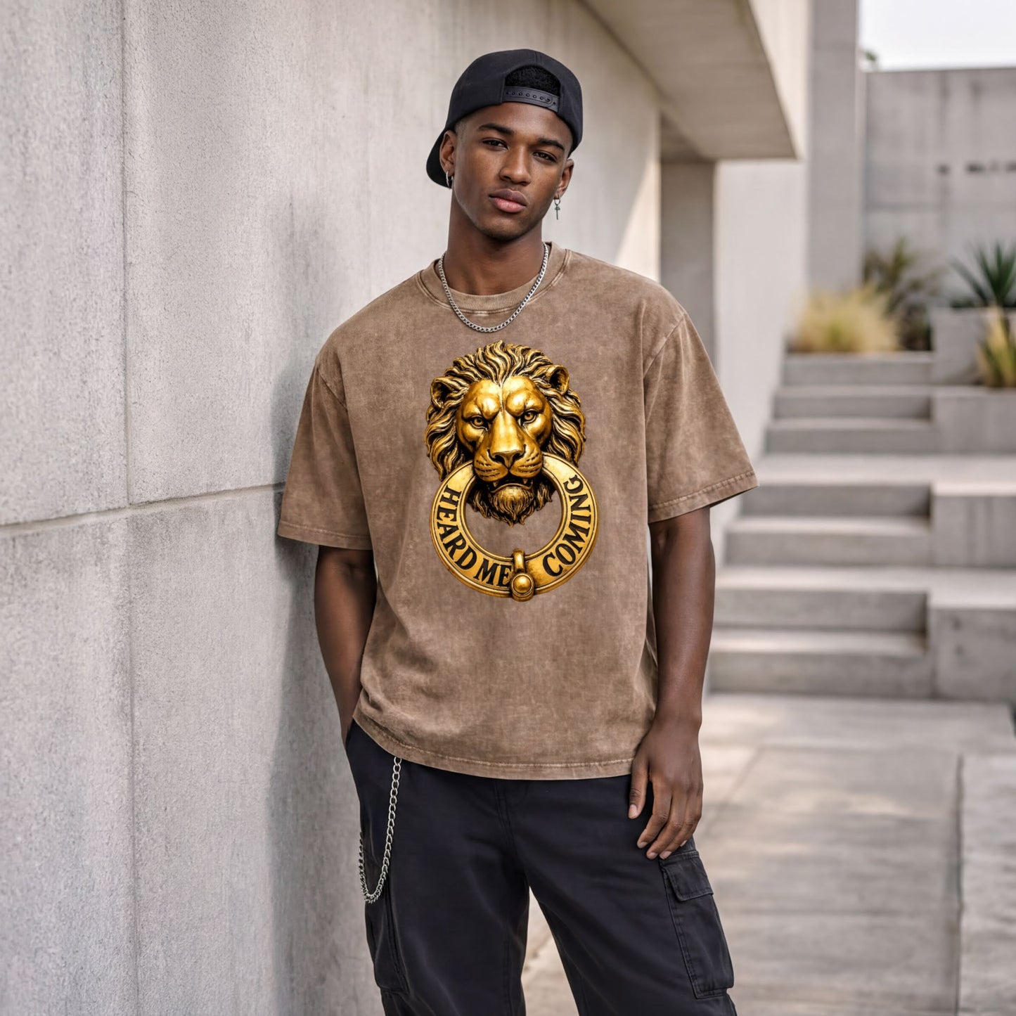 A young man wearing a brown t-shirt with a lion's head design and the text "HEARD ME COMING" stands in front of a concrete wall.
