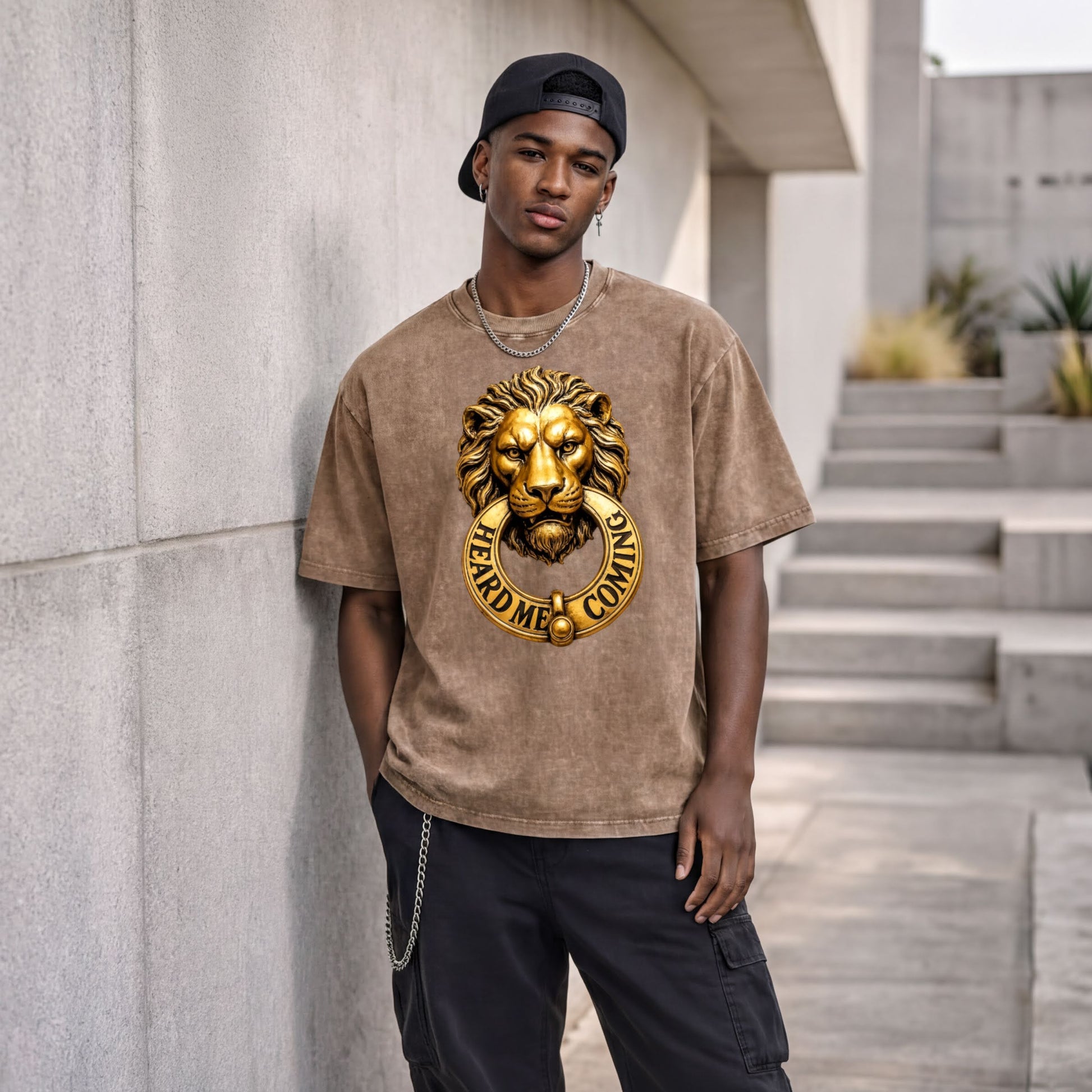 A young man wearing a brown t-shirt with a lion's head design and the text "HEARD ME COMING" stands in front of a concrete wall.