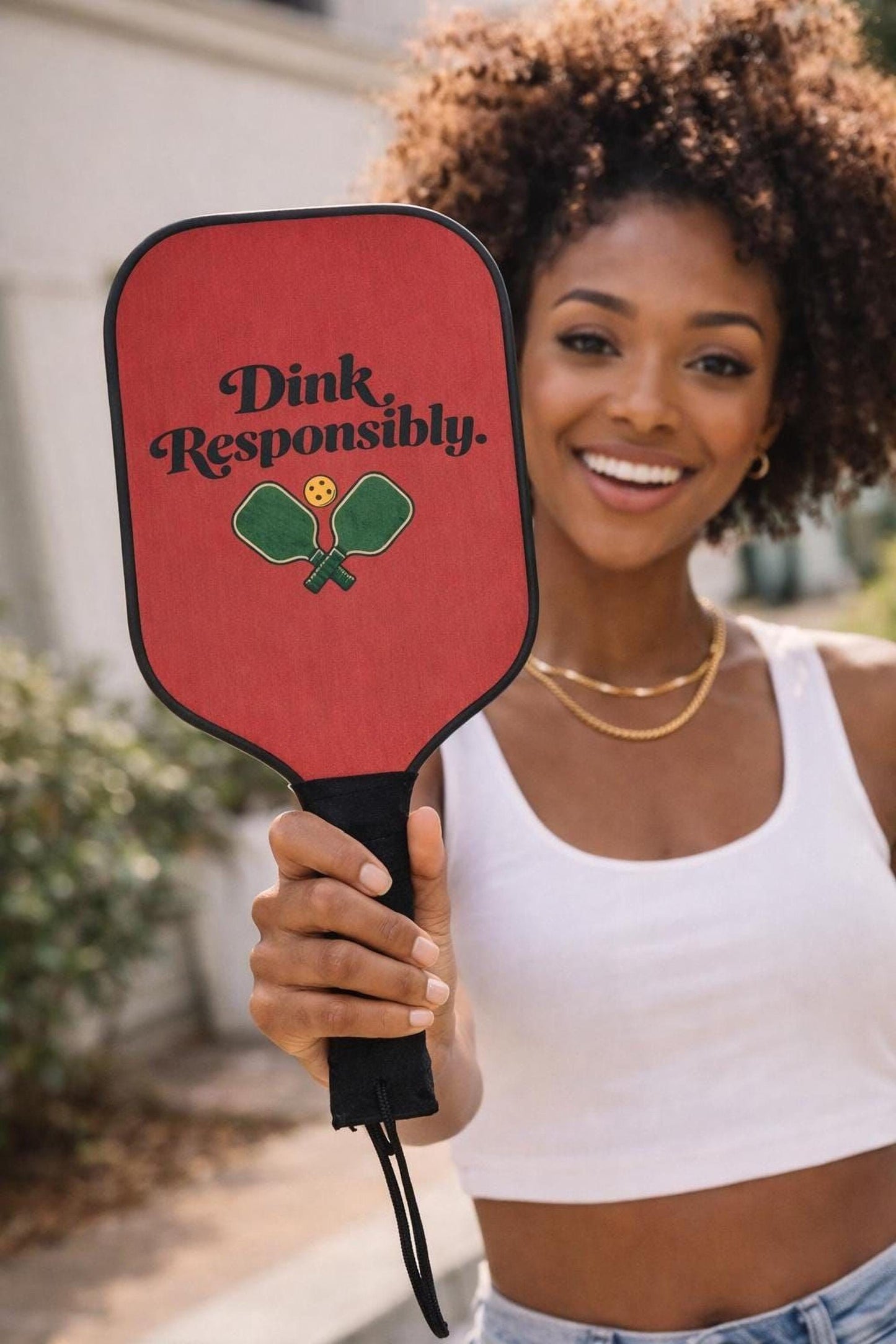 A woman with curly hair is holding a red paddle with the text "Dink Responsibly" printed on it.