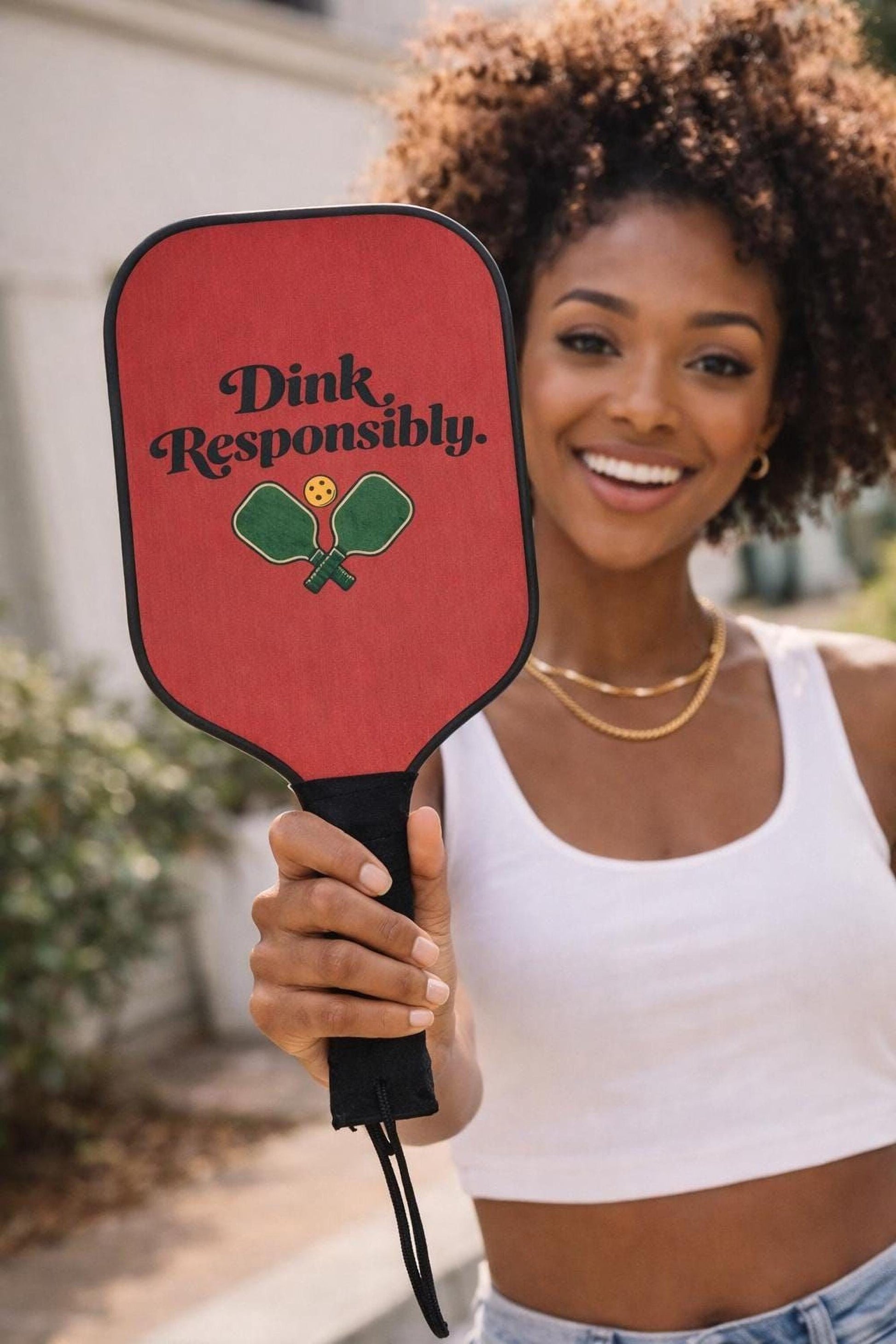 A woman with curly hair is holding a red paddle with the text "Dink Responsibly" printed on it.