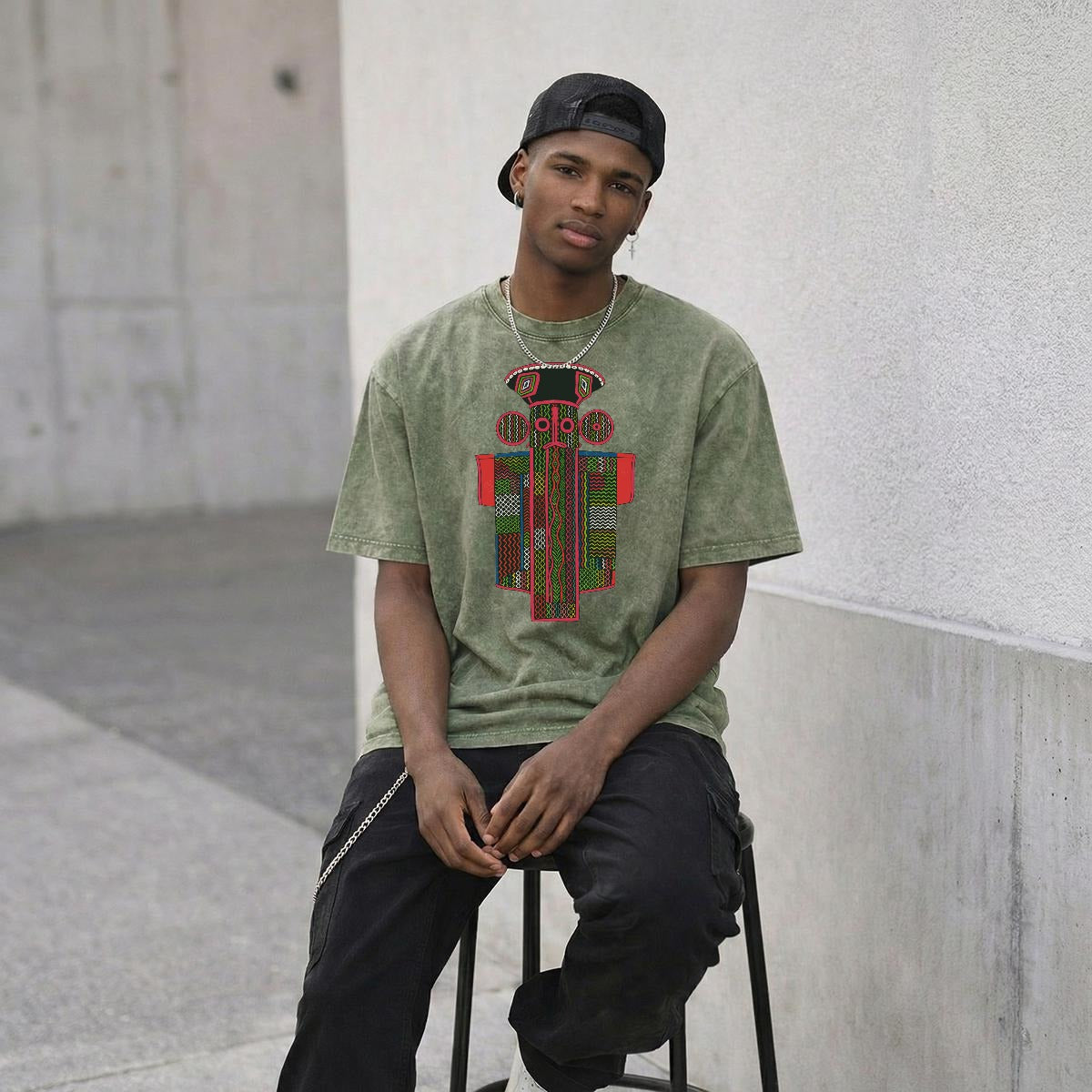 A young man wearing a green t-shirt with a colorful graphic design, sitting on a stool and looking directly at the camera.