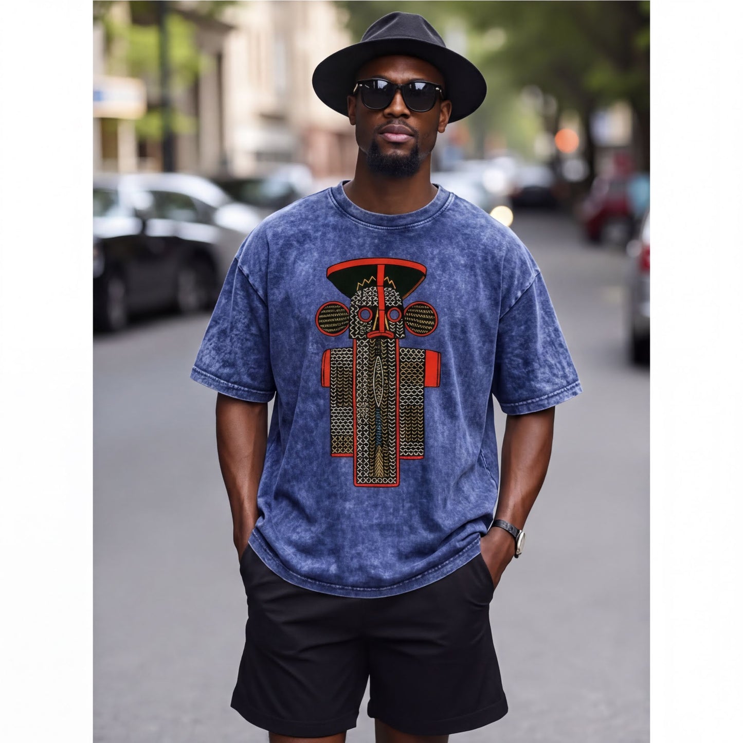 A man wearing a blue t-shirt with a graphic design, black shorts, and a black hat stands on a city street.