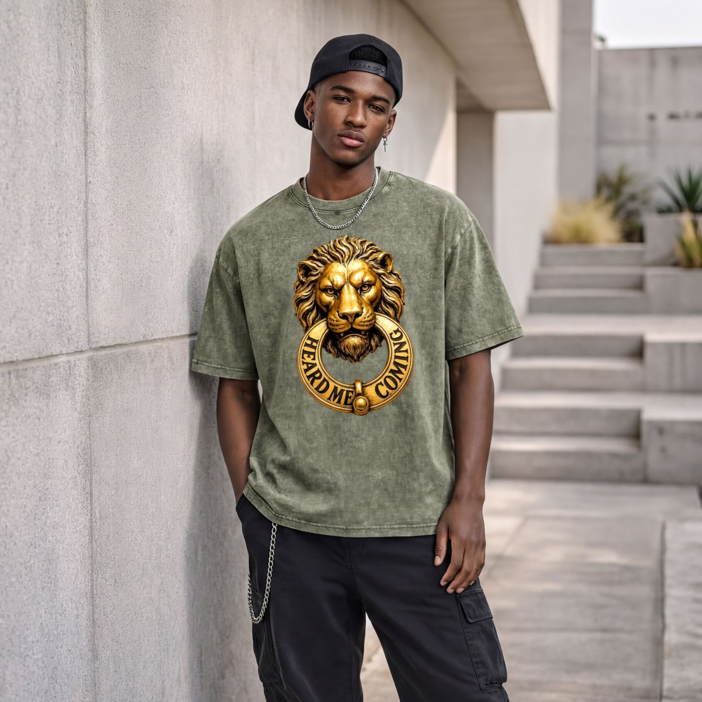 A young man wearing a green t-shirt with a lion's head design and a chain around his neck is standing against a concrete wall.