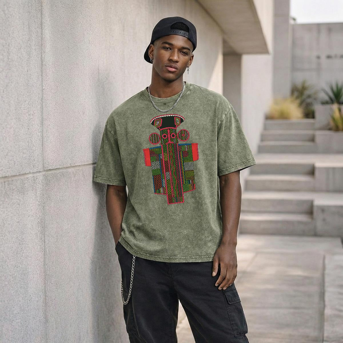 A young man wearing a green t-shirt with a graphic design stands in front of a concrete wall, leaning against it.