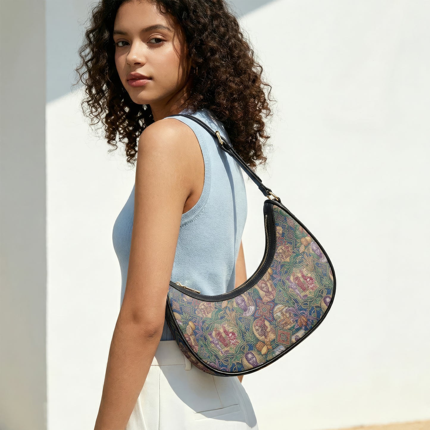 A young woman with curly hair is standing against a white wall, wearing a blue sleeveless top and carrying a colorful, patterned purse.