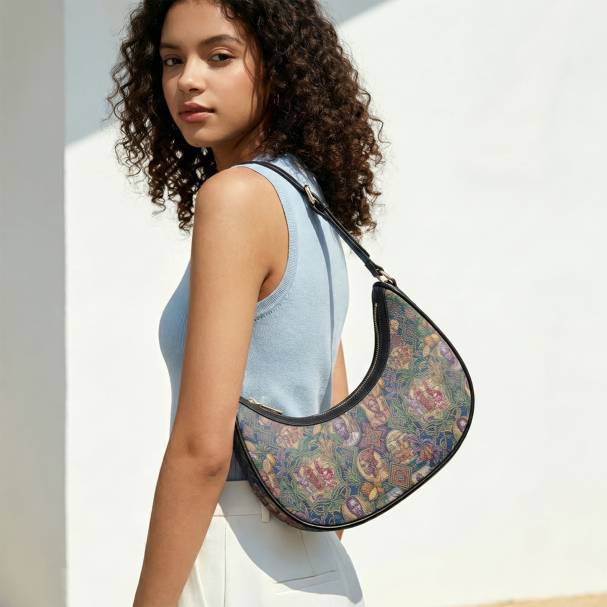 A young woman with curly hair is standing against a white wall, wearing a blue sleeveless top and carrying a colorful, patterned purse.