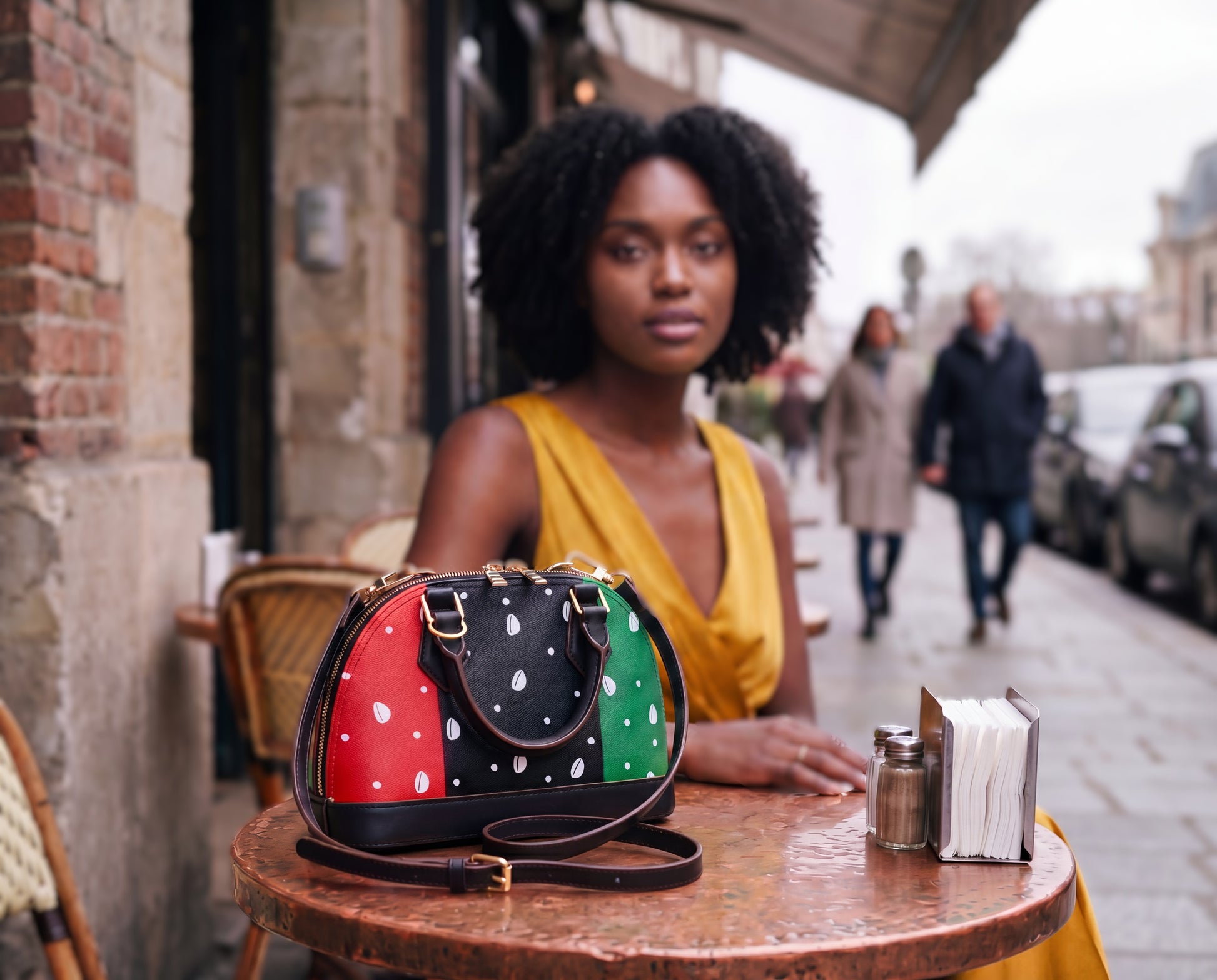 A woman in a yellow dress sits at a table with a polka dot handbag in front of her, with a city street and other people visible in the background.