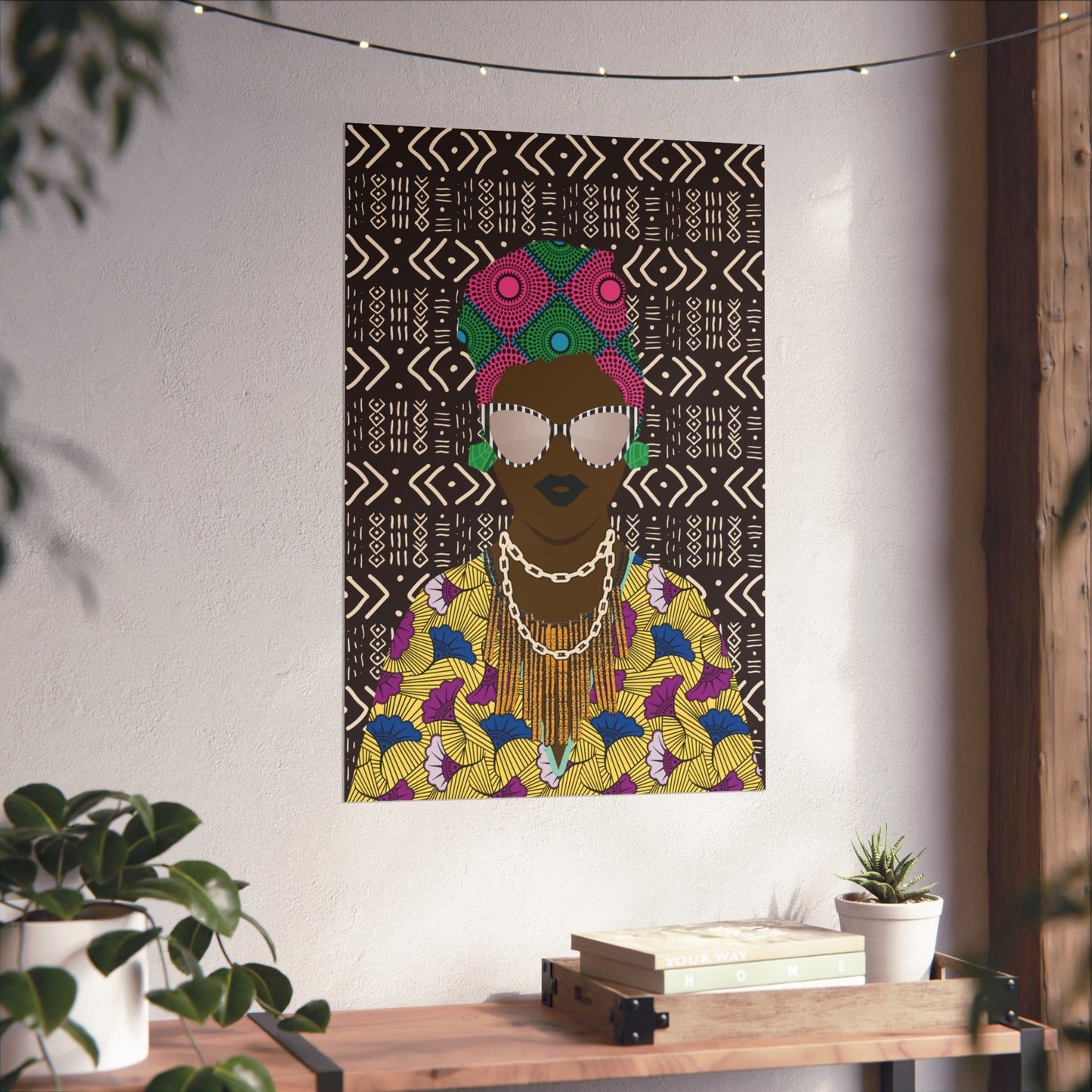a colorful poster of a woman with a headdress hanging on a wall, surrounded by a potted plant and a wooden table with books on it.