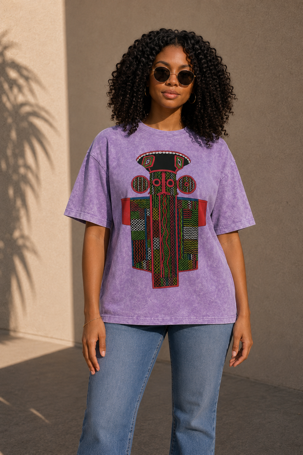 A woman with curly hair wearing a purple t-shirt with a colorful graphic design stands in front of a beige wall.