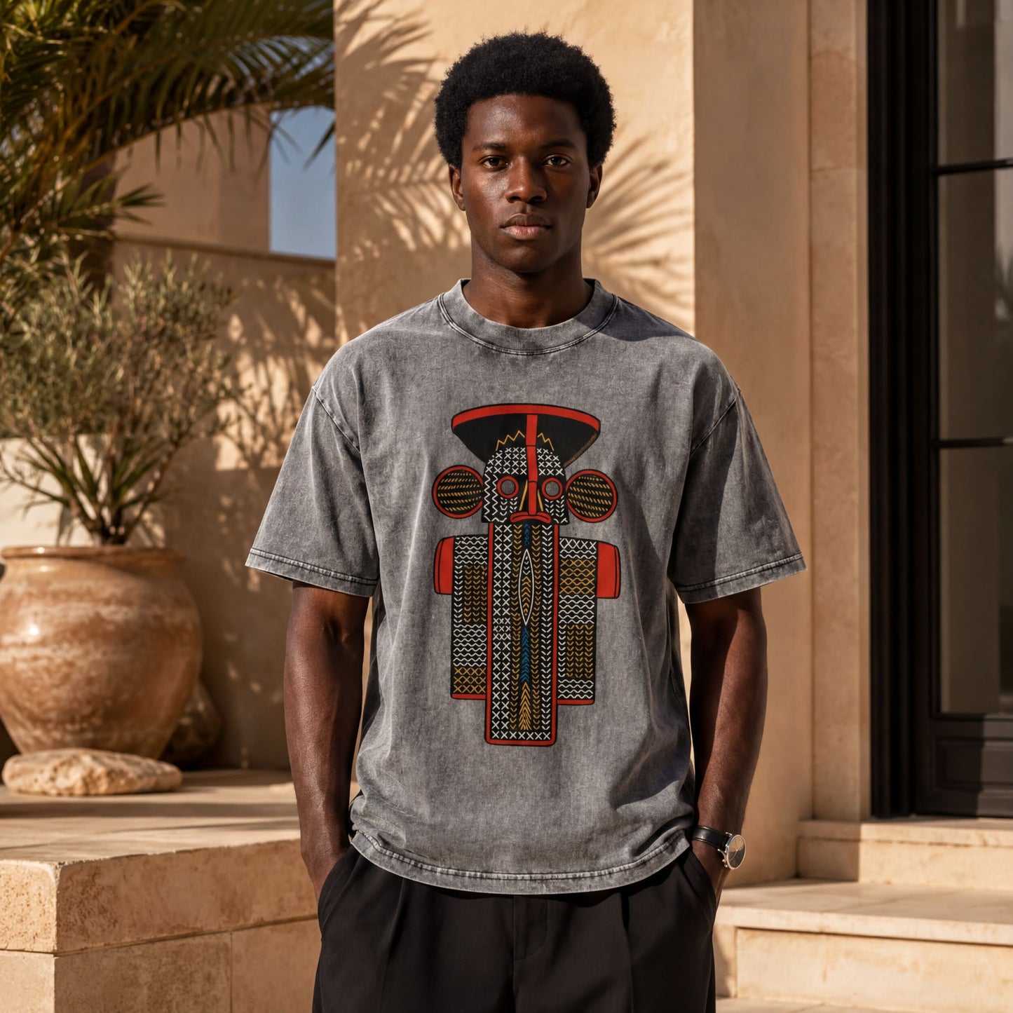 A young man wearing a gray t-shirt with a graphic design stands in front of a building with palm trees in the background.