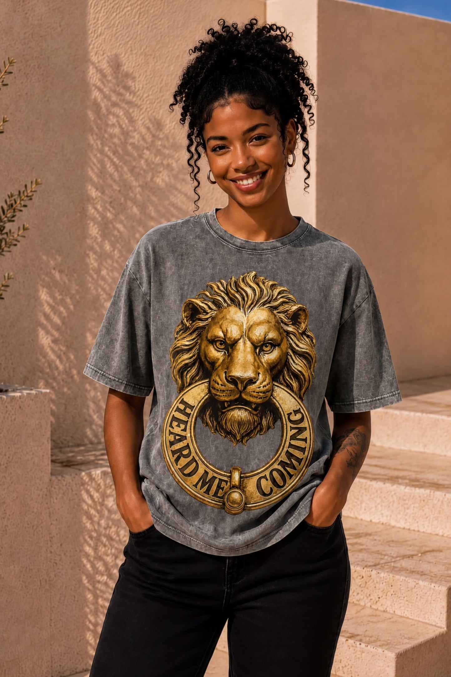 A smiling woman wearing a gray t-shirt with a lion's head design and the text "HEARD ME COMING" printed on it.