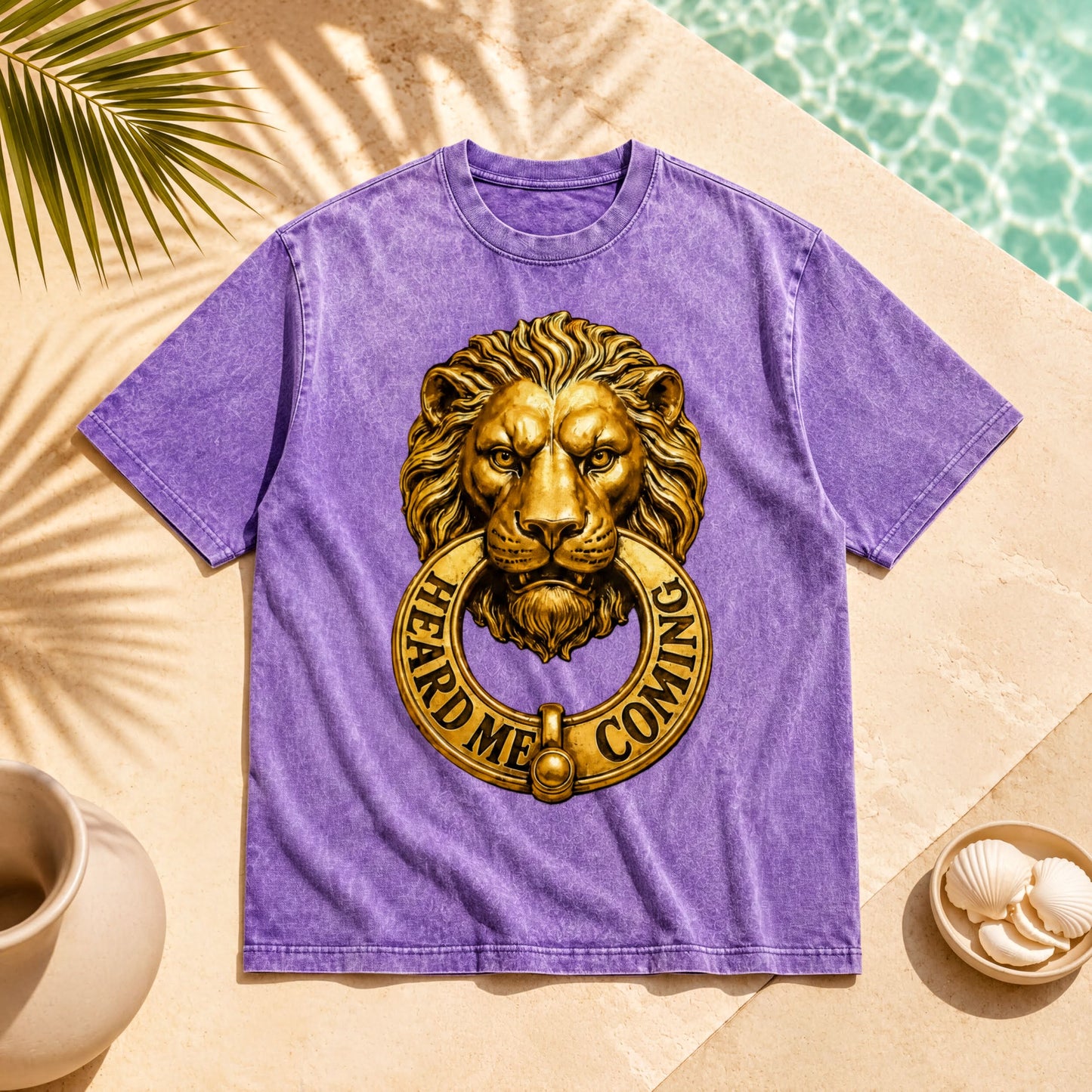 a purple t-shirt with a lion's head design and the text "HEARD ME COMING" printed on it. The t-shirt is placed on a beige surface, with a palm frond and a small bowl of shells nearby.