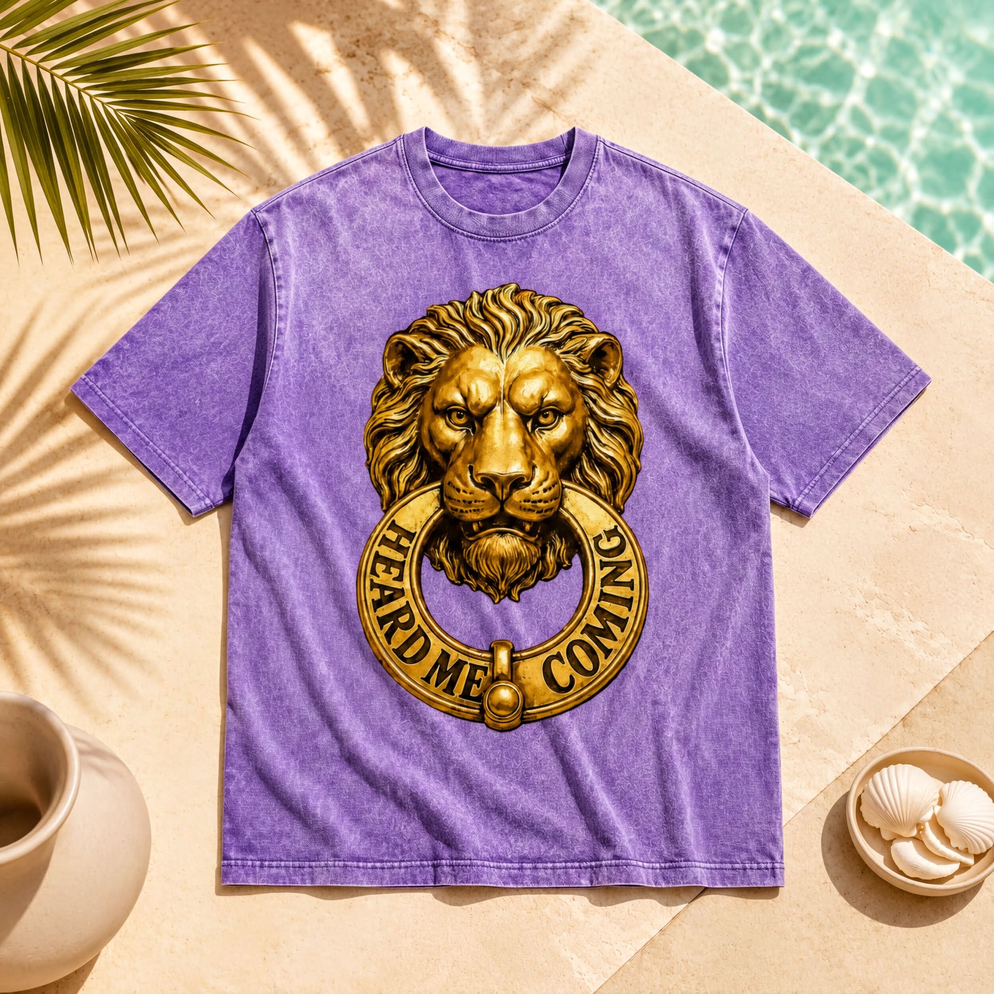 a purple t-shirt with a lion's head design and the text "HEARD ME COMING" printed on it. The t-shirt is placed on a beige surface, with a palm frond and a small bowl of shells nearby.