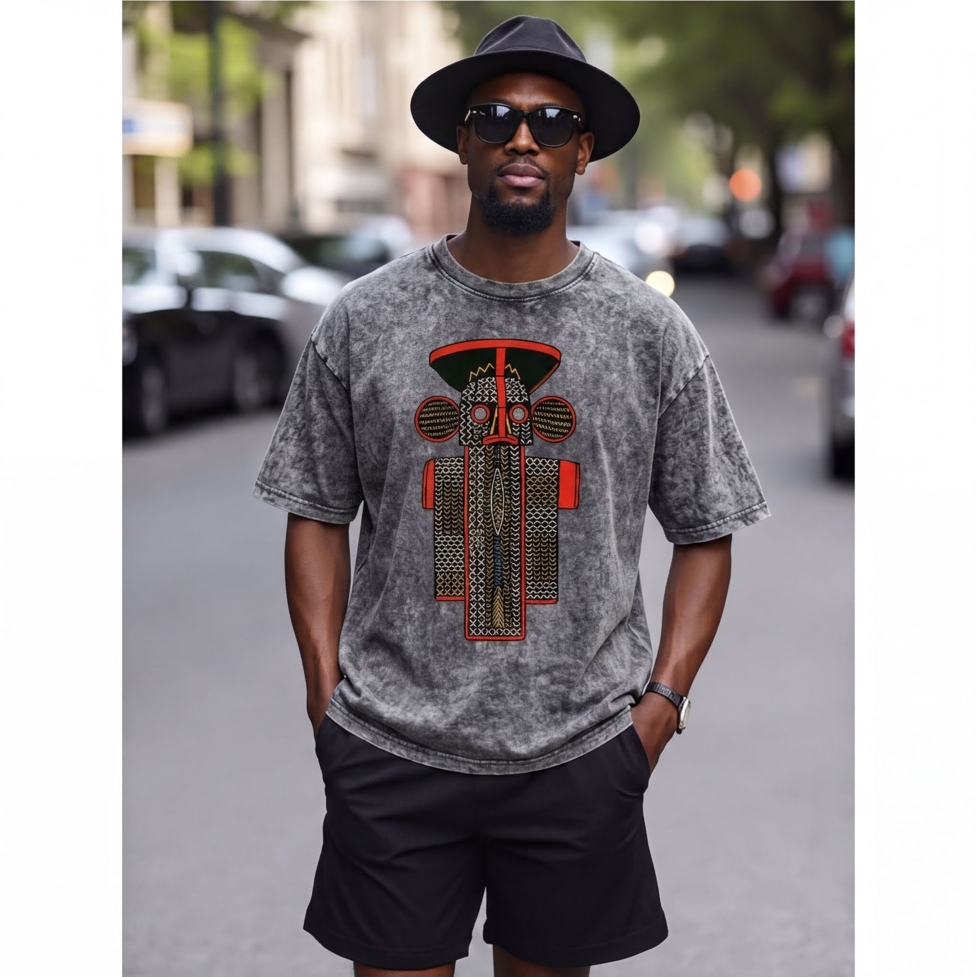 A man wearing a gray t-shirt with a graphic design, black shorts, and a black hat is standing on a city street.