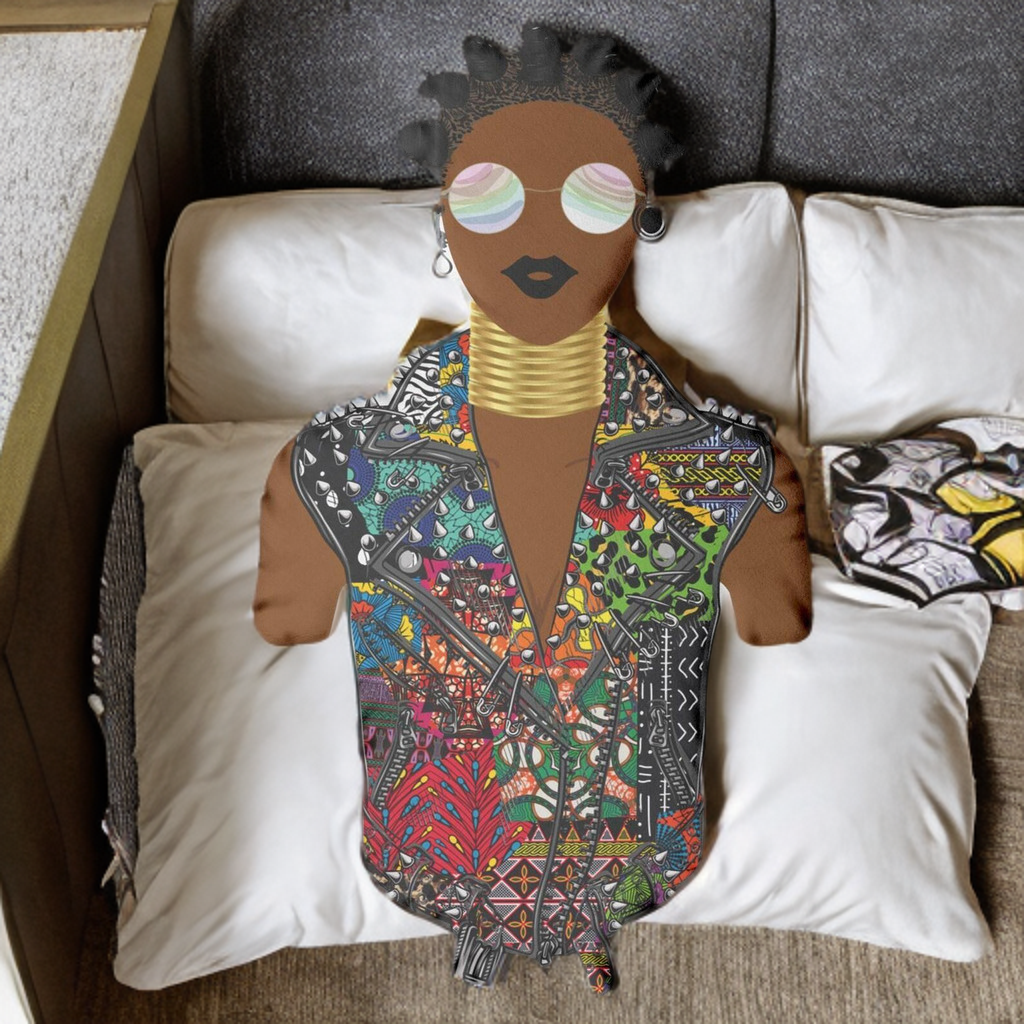 A colorful, patterned doll with a unique hairstyle and large sunglasses sits on a bed surrounded by pillows.