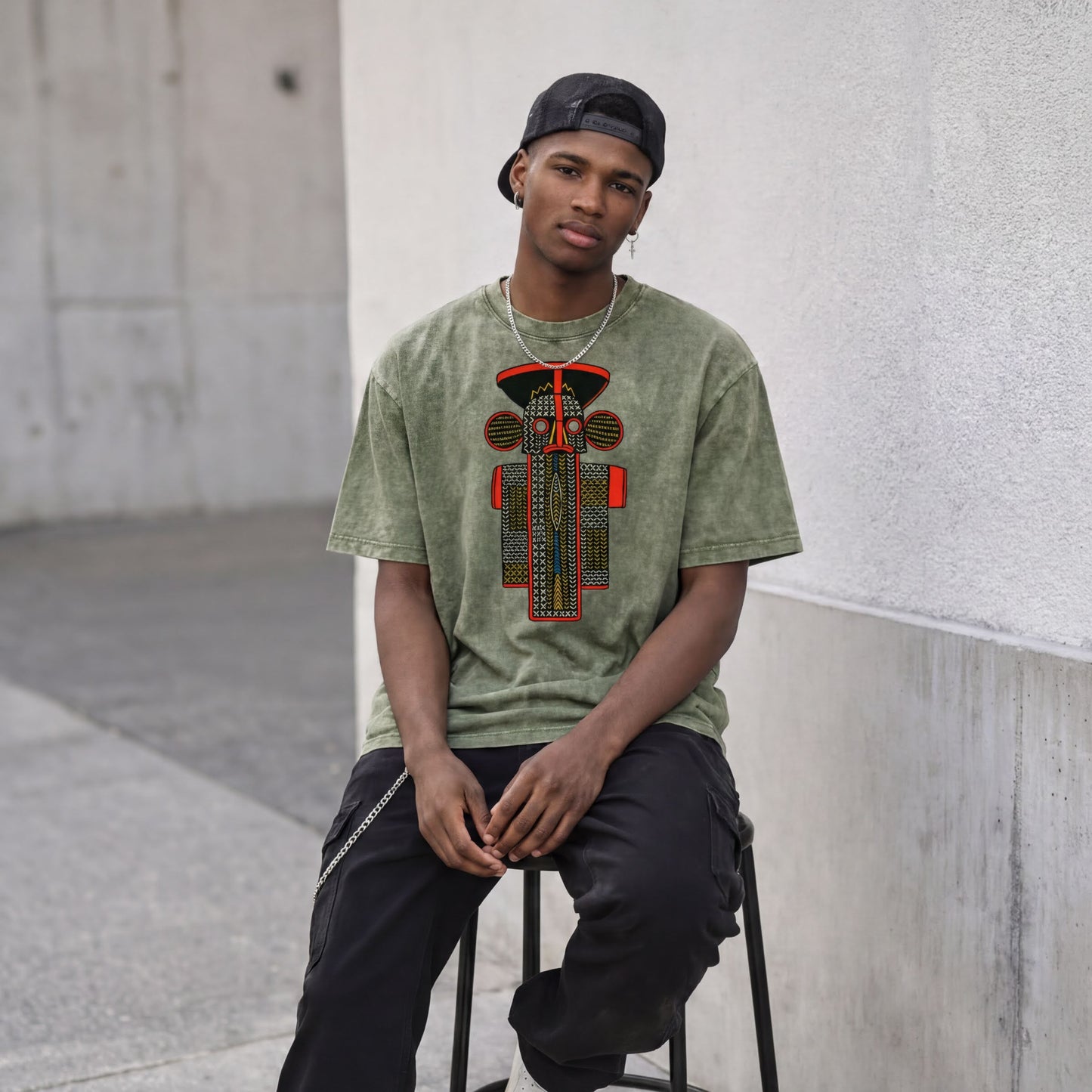 A young man wearing a green t-shirt with a graphic design and a black cap is sitting on a stool against a concrete wall.