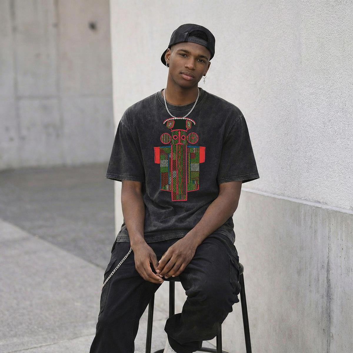 A young man wearing a black t-shirt with a colorful graphic and a black cap is sitting on a stool against a concrete wall.