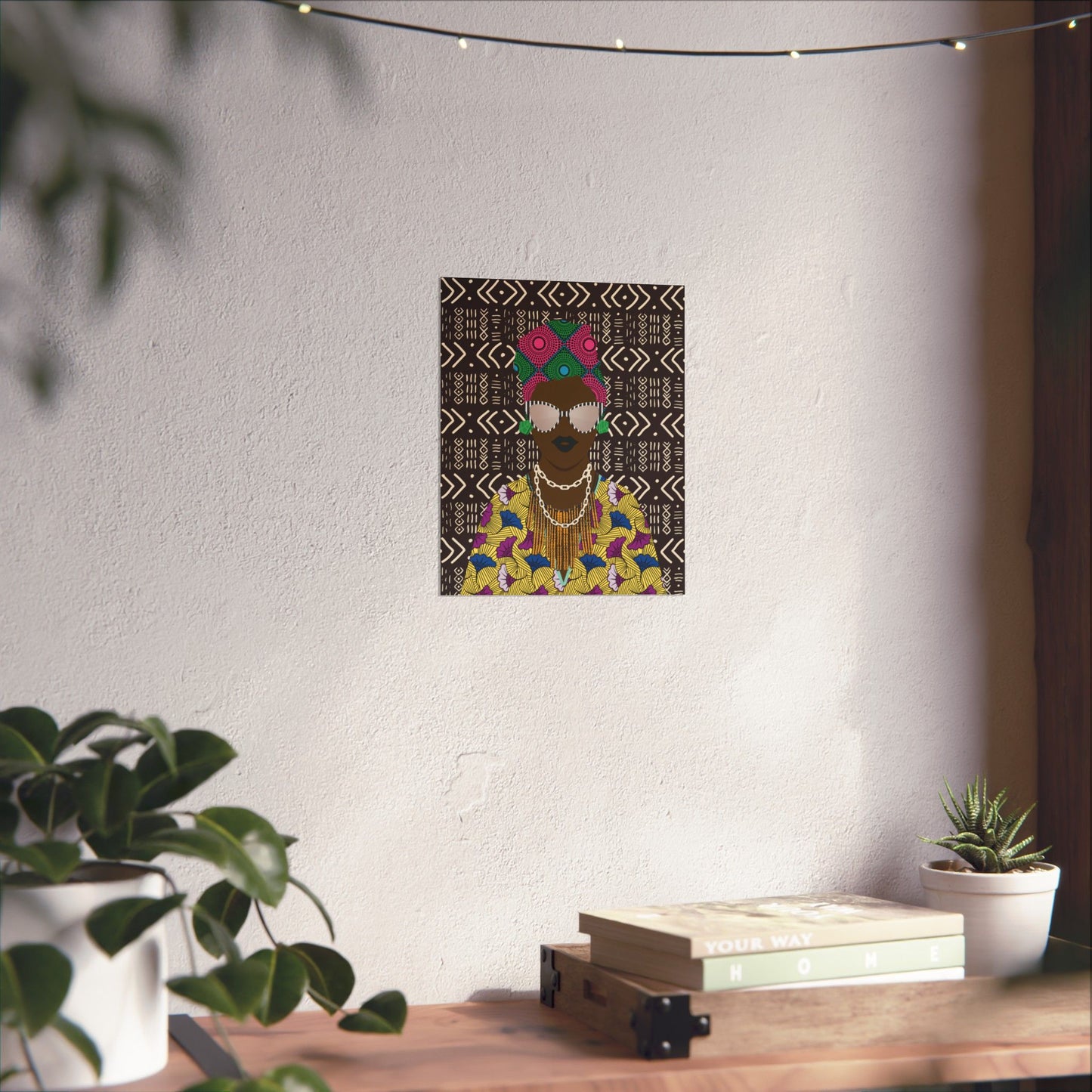 a wall with a framed artwork depicting a person wearing a colorful, patterned outfit and a headpiece. The artwork is hanging on the wall, and there are potted plants and a stack of books on a table nearby.