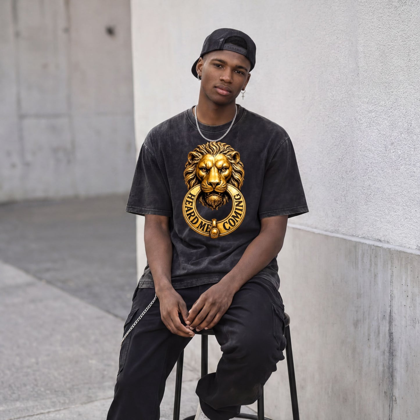 A young man wearing a black t-shirt with a lion head design and a chain necklace is sitting on a stool against a white wall.
