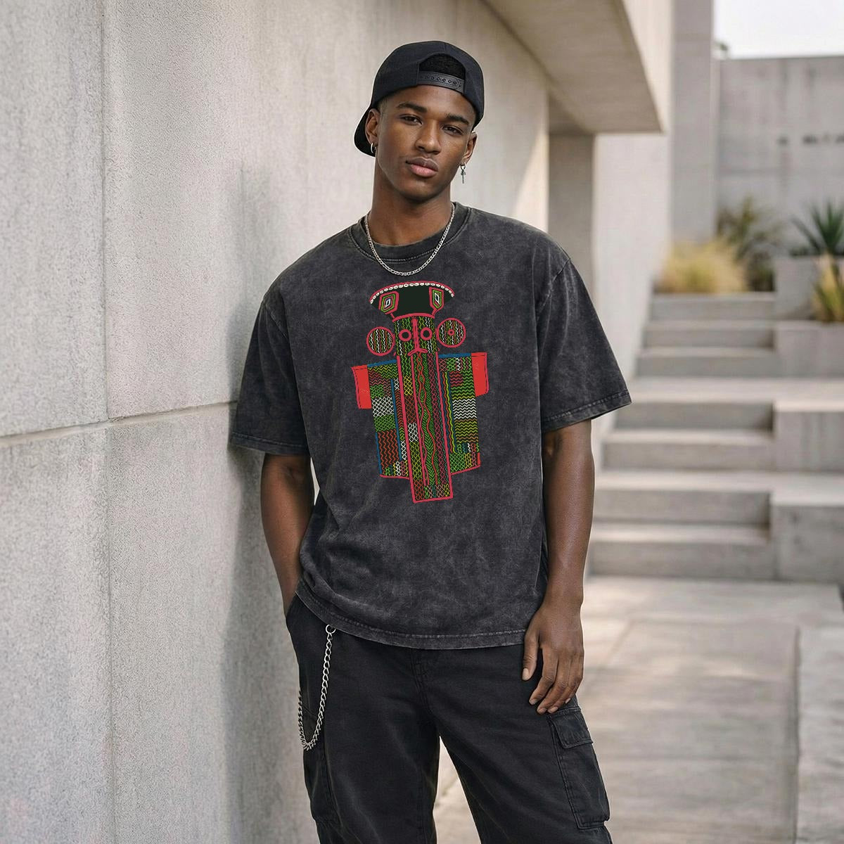 A young man wearing a black t-shirt with a graphic design stands in front of a concrete wall, leaning against it.