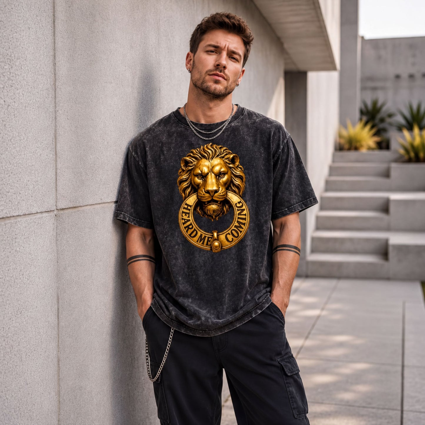A man wearing a black t-shirt with a lion head design stands against a gray wall, with a chain around his neck.
