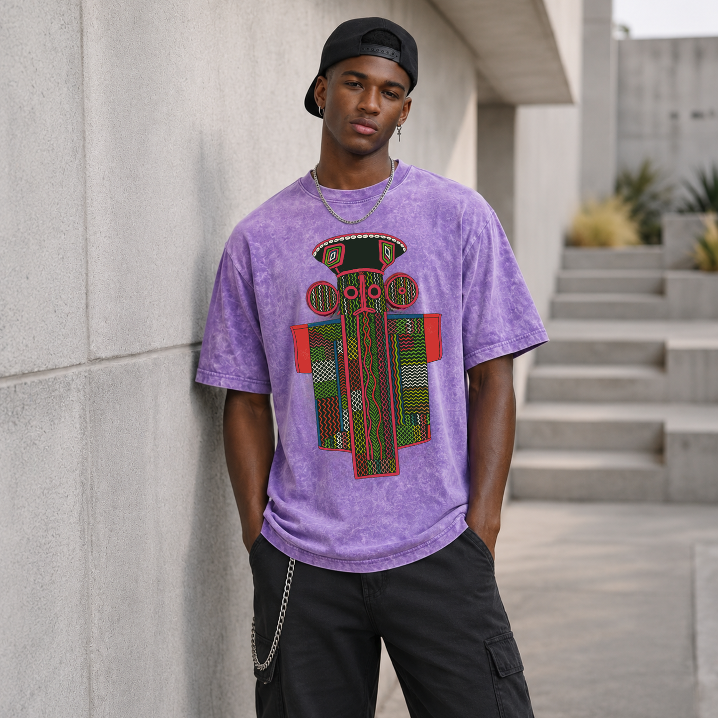 A young man wearing a purple t-shirt with a colorful graphic design stands in front of a concrete wall, with a staircase visible in the background.