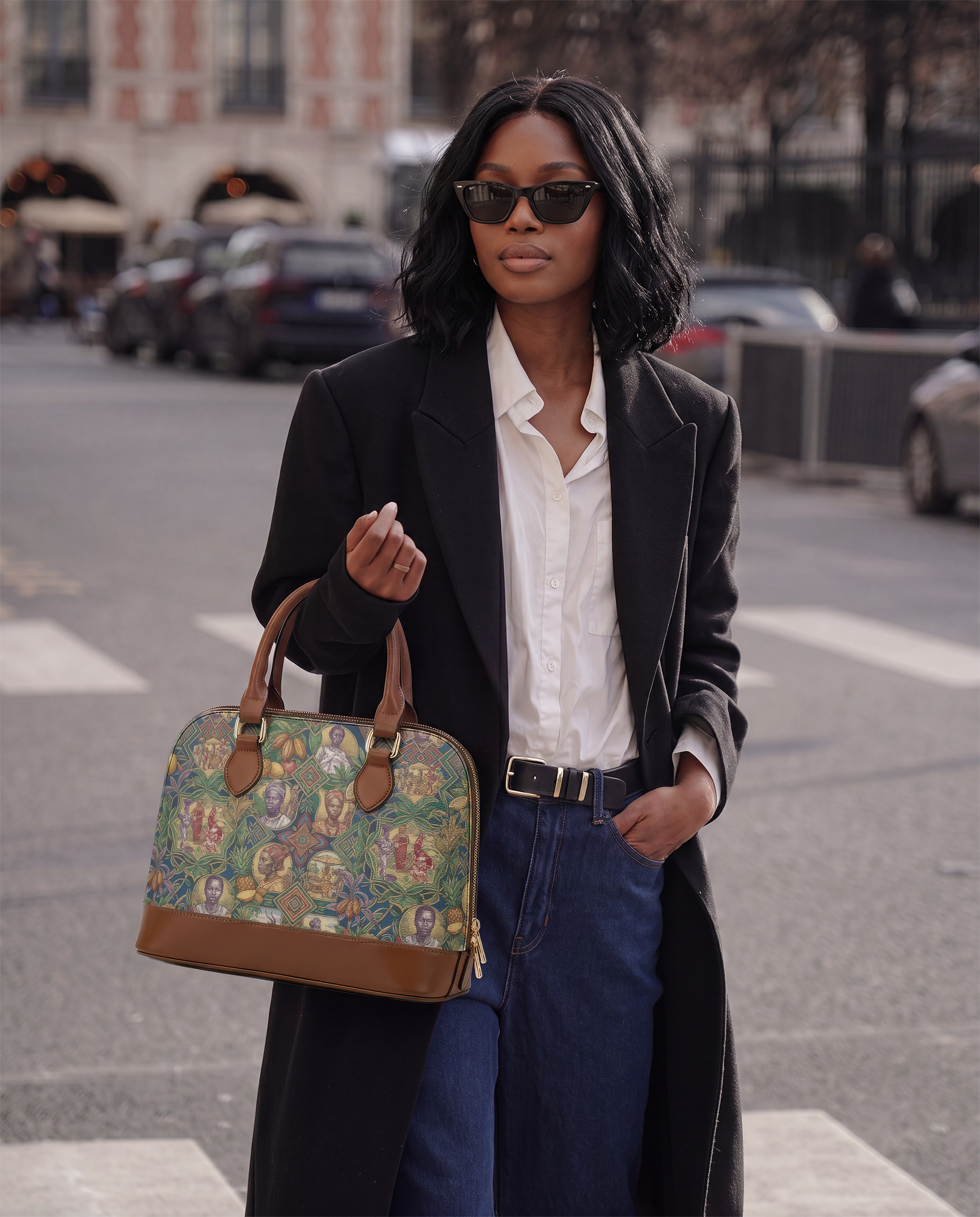 A woman wearing a black coat, white shirt, and sunglasses is walking down a city street, carrying a colorful handbag.