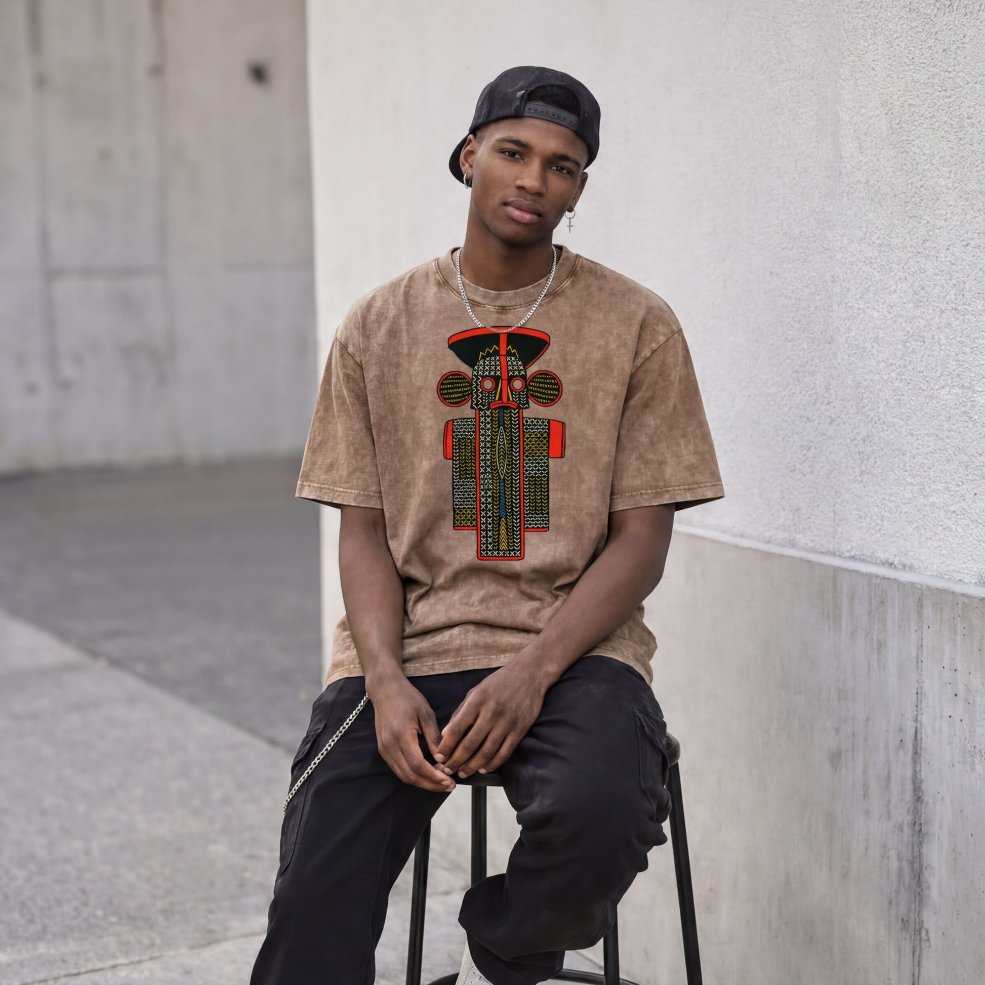 A young man wearing a brown t-shirt with a graphic design and a black cap is sitting on a stool against a white wall.