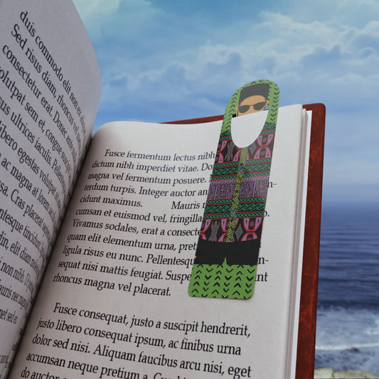 May include: A green bookmark features a stylized figure wearing sunglasses and an ethnic-patterned outfit. The bookmark has a rectangular shape with a pointed bottom.  The figure's outfit includes black pants and a green, black, and pink patterned top. The bookmark is shown inserted into a book with text visible, including Latin phrases. The background shows a blurred ocean view.