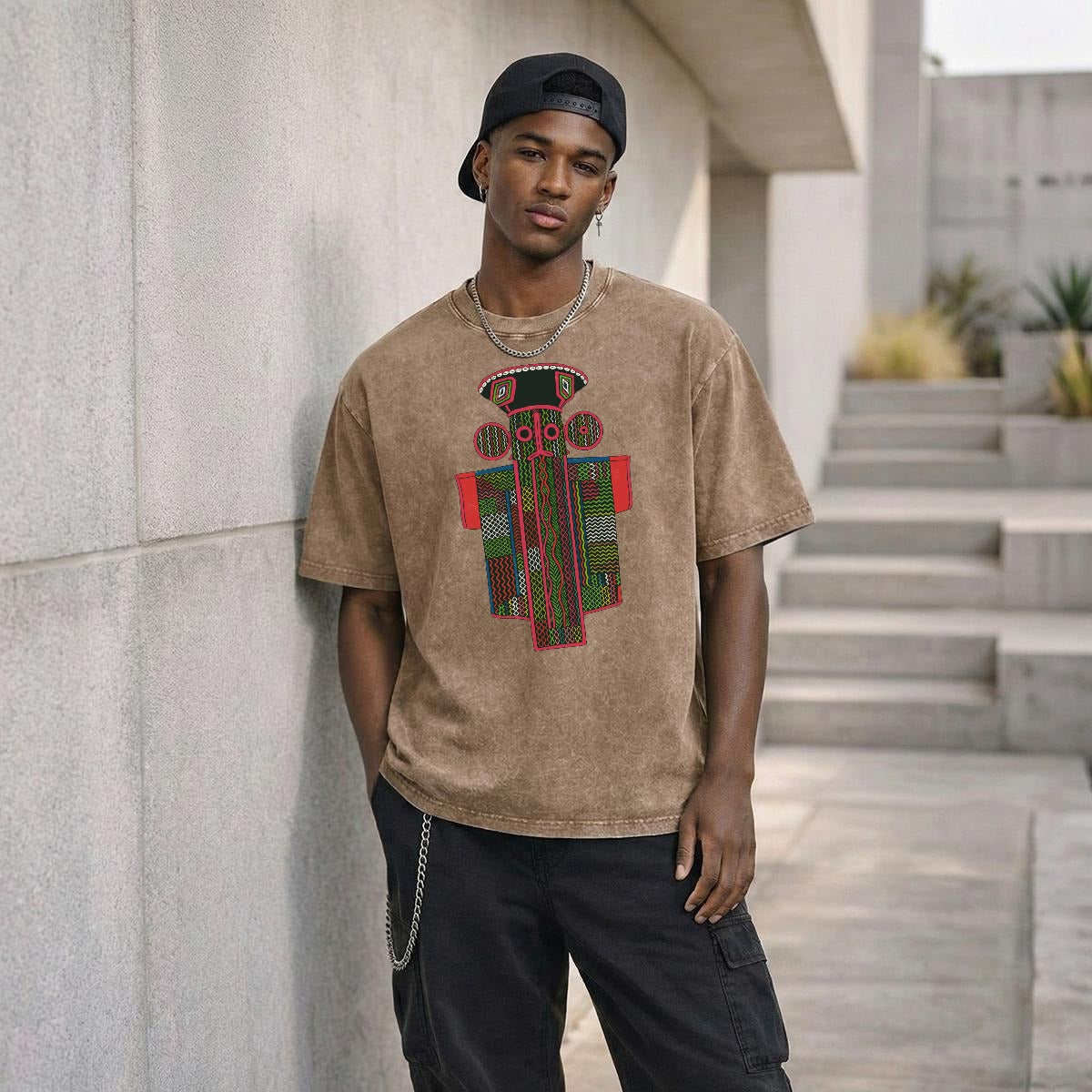 A young man wearing a brown t-shirt with a colorful graphic design stands in front of a concrete wall, leaning against it.