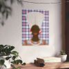 a wall with a framed artwork depicting a person's head wearing a hat, surrounded by a checkered pattern. The artwork is hanging on a white wall, and there are two potted plants and a stack of books placed on a wooden surface below it.