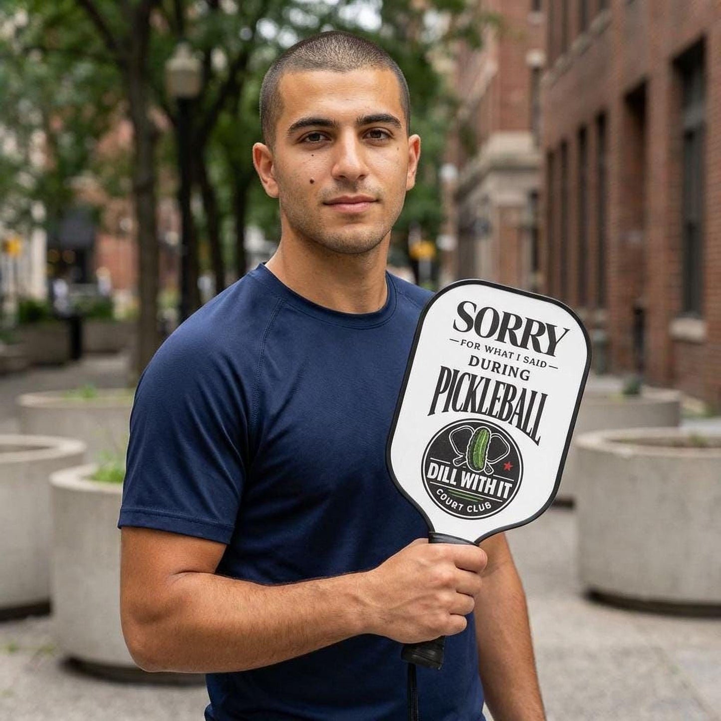 A man in a blue shirt is holding a white paddle with a humorous message on it, standing in front of a brick building.