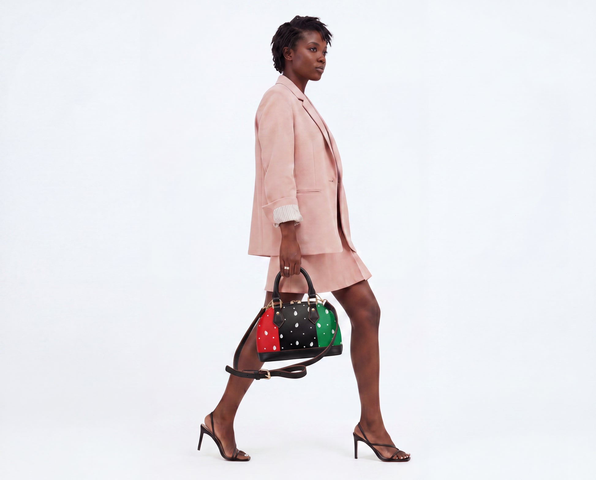 A woman in a pink suit and high heels is walking and carrying a colorful handbag with polka dots.
