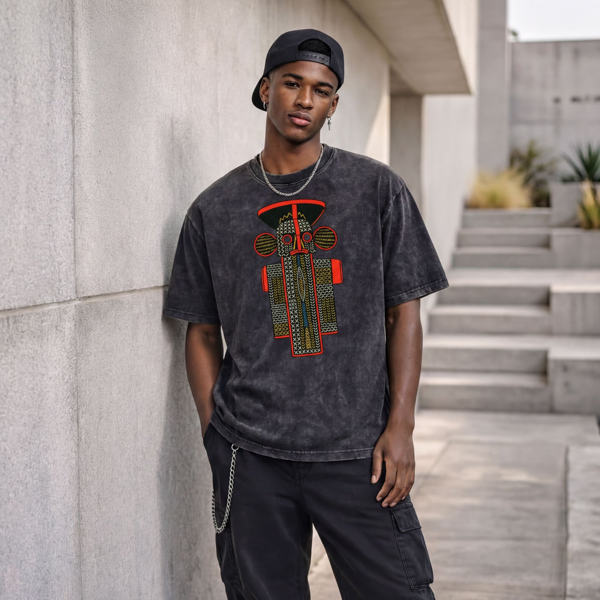 A young man wearing a black t-shirt with a graphic design stands against a concrete wall, with a chain hanging from his pocket.