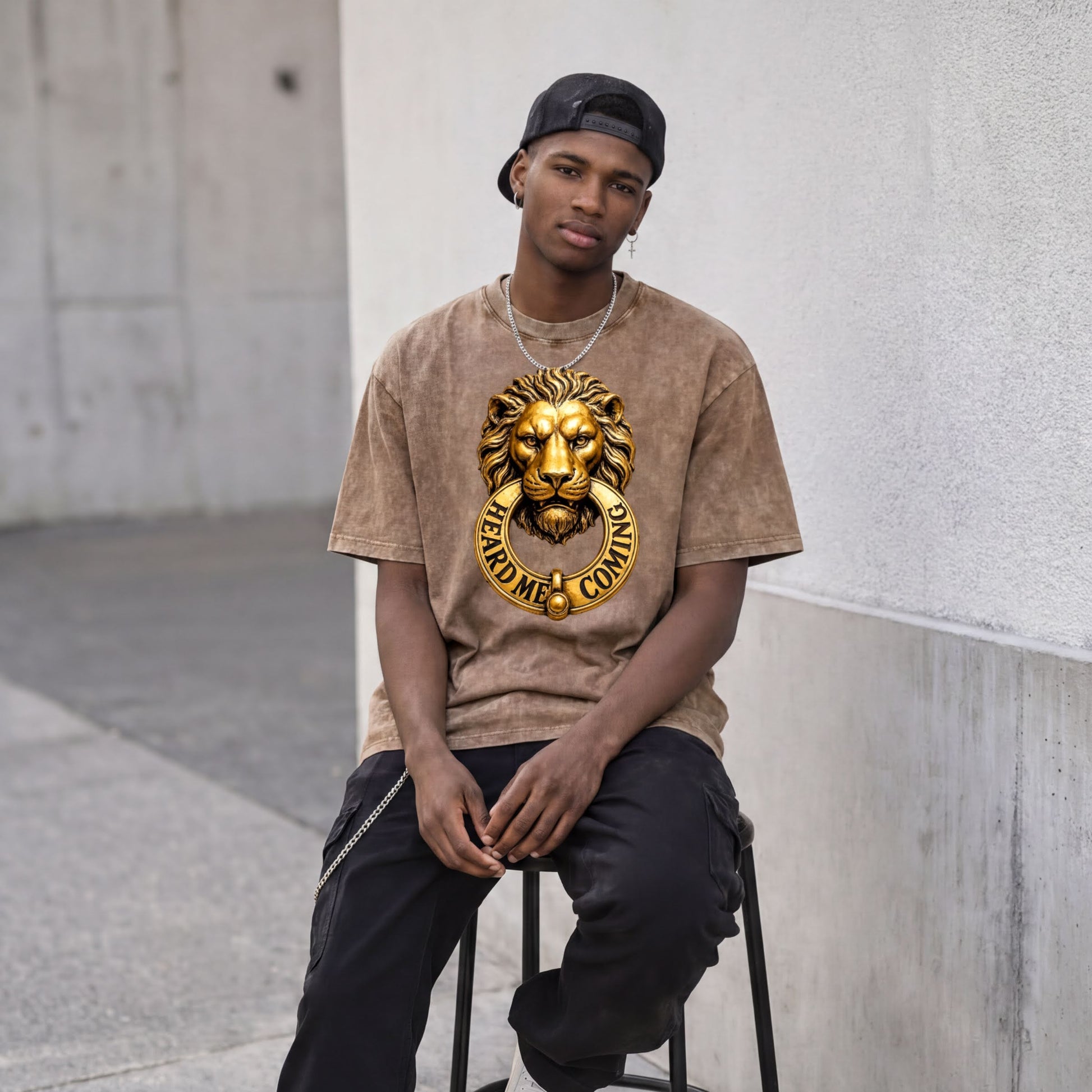 A young man wearing a brown t-shirt with a lion head graphic and the text "Hear me, coming" is sitting on a stool against a concrete wall.