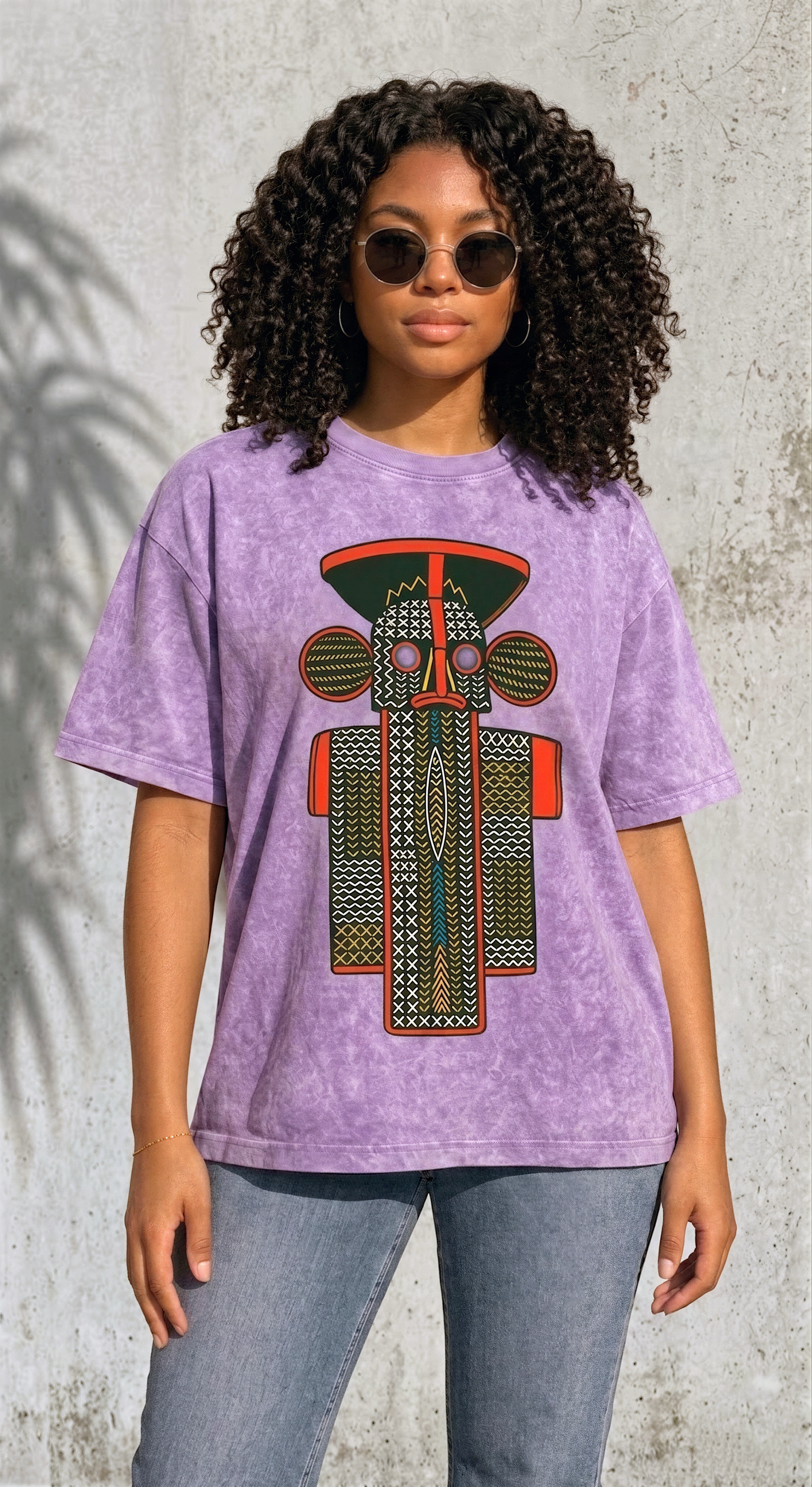 A young woman with curly hair wearing a purple t-shirt with a colorful graphic design stands in front of a concrete wall.