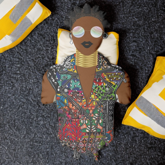 A colorful, patterned doll with a unique hairstyle and large sunglasses, sitting on a gray carpet with yellow and white pillows in the background.