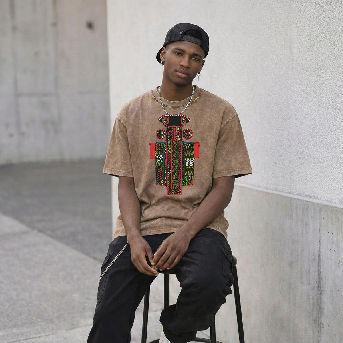 A young man wearing a beige t-shirt with a colorful graphic design, sitting on a stool and looking directly at the camera.