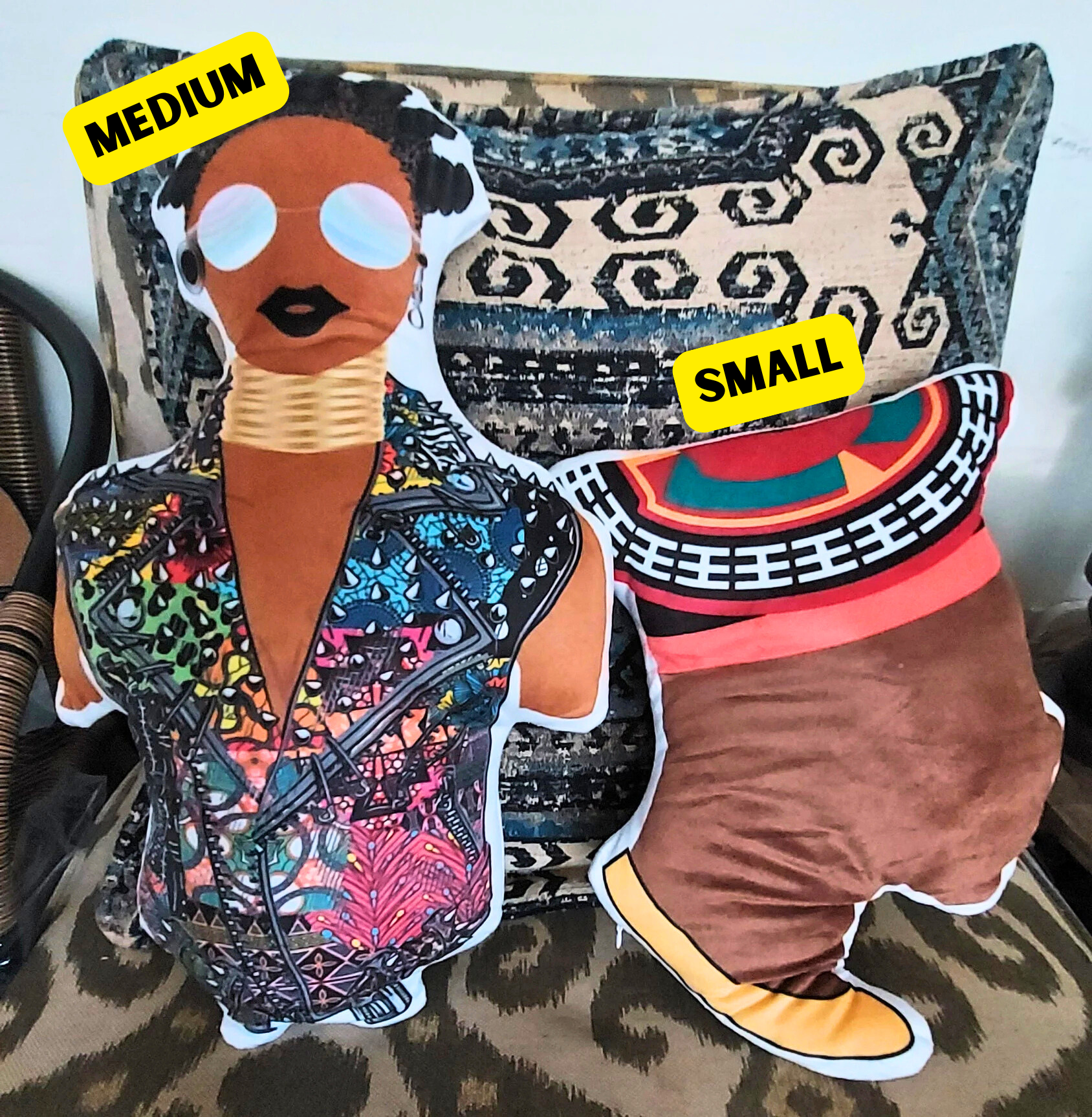 two stuffed animal figures, one with a colorful patterned outfit and the other with a brown outfit, placed on a patterned cushion.