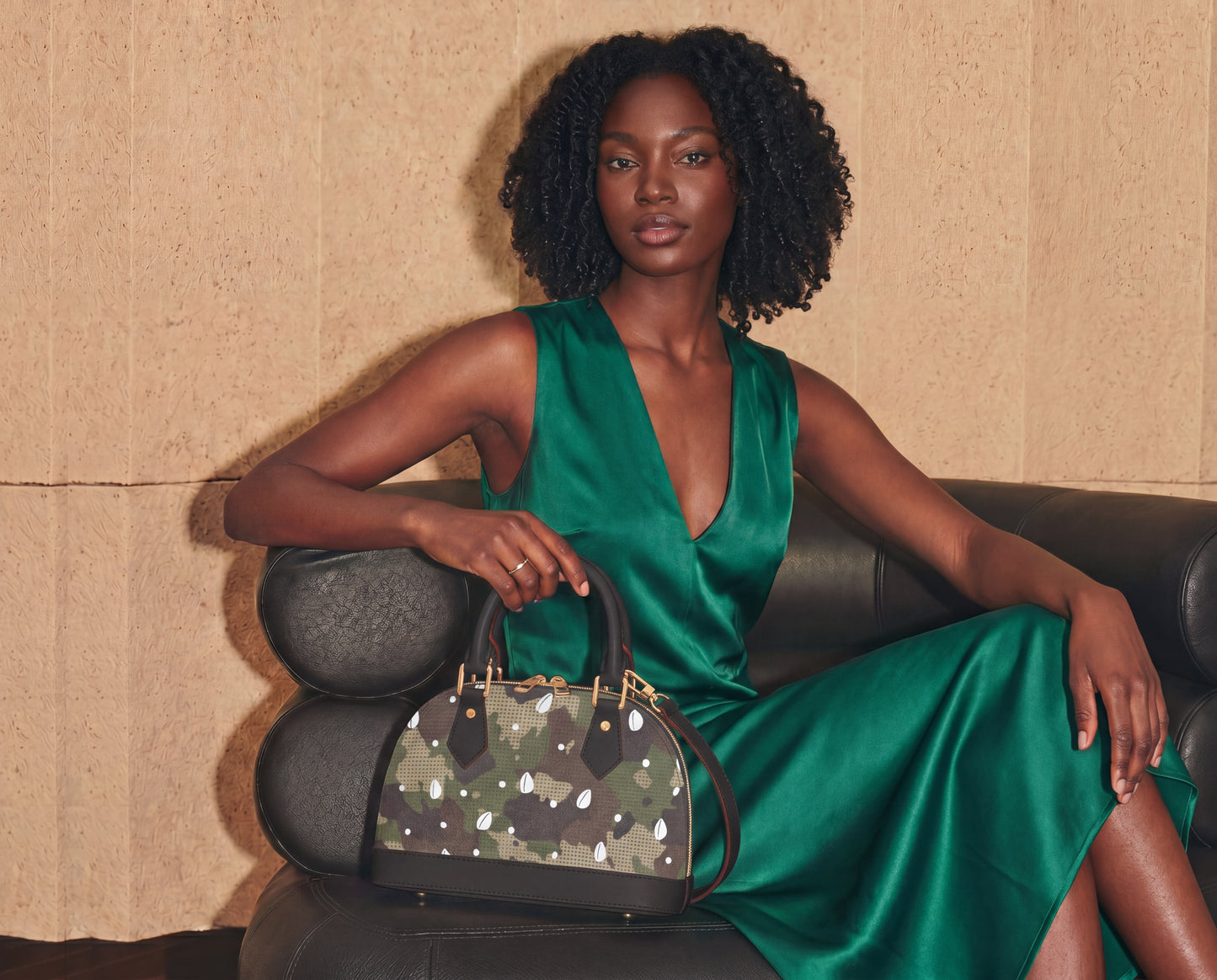 A woman in a green dress sits on a black leather couch, holding a black and gray handbag with a camouflage pattern.